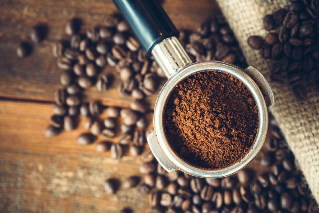 Freshly ground espresso in a portafilter, ready to be made into a hot shot of espresso.  Rustic wood background, a burlap coffee bag in the background covered with roasted coffee beans.  Overhead view.